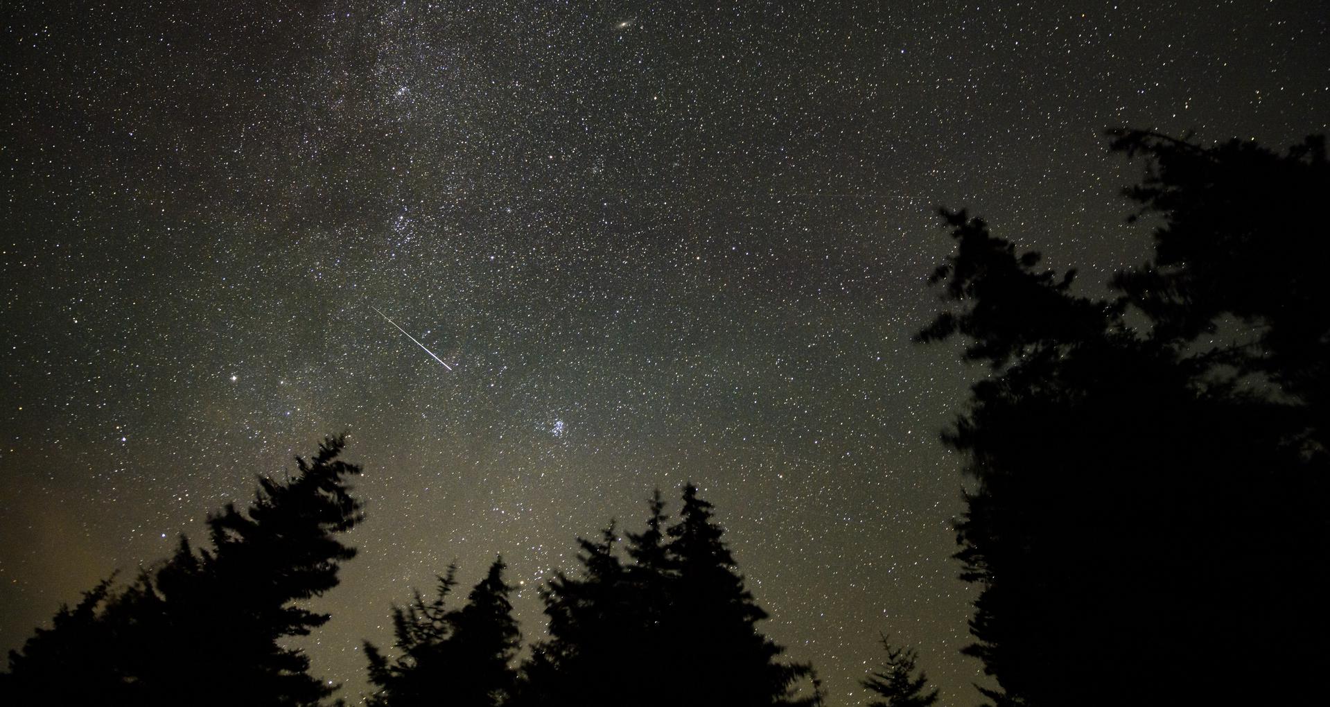 Un météore traverse le ciel étoilé au-dessus d'un horizon sombre, la Voie lactée en fond